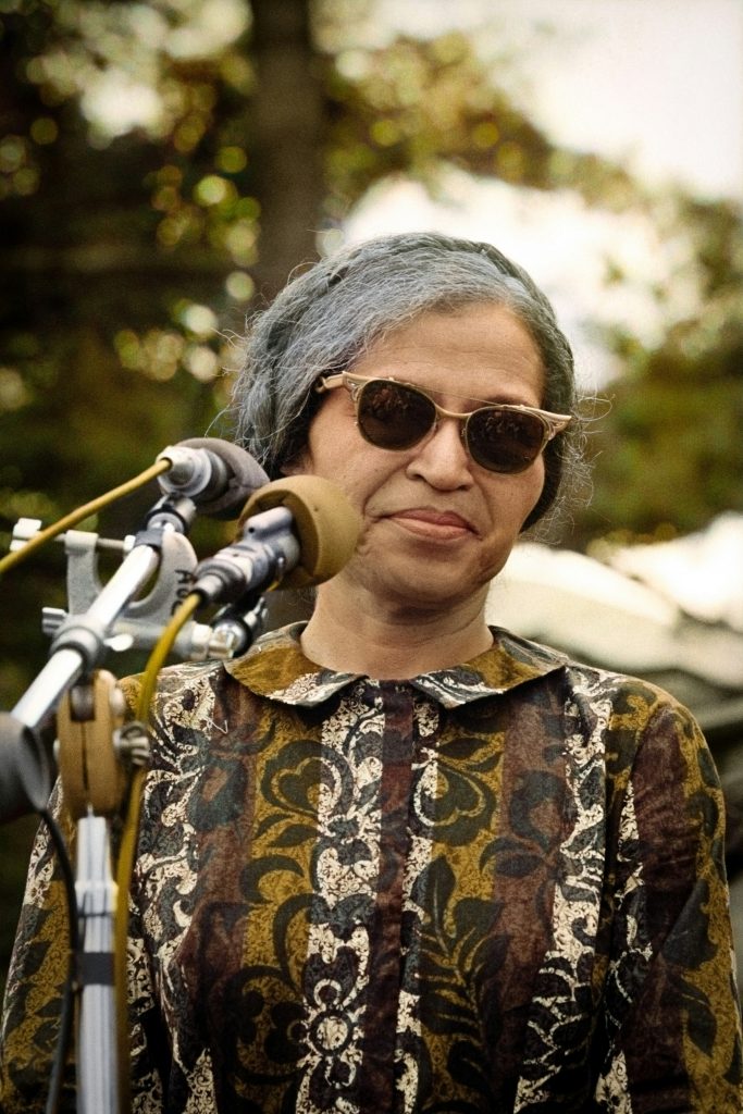 Photo of Rosa Parks wearing sunglasses standing in front of a microphone